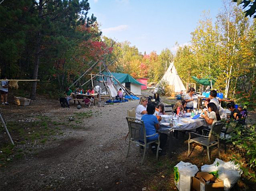 group of people of various ages eating together outside on tables with teepee in background